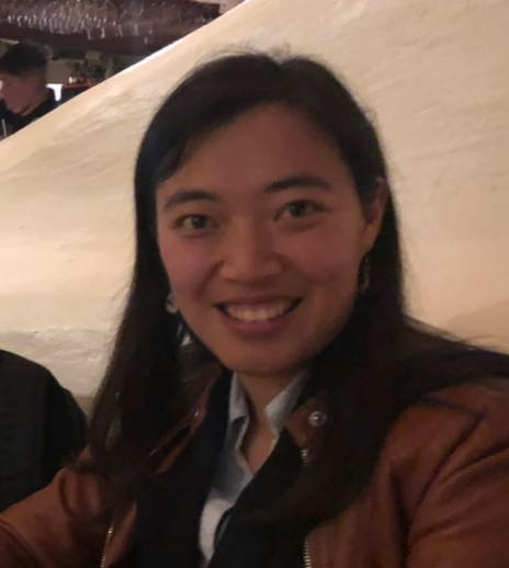 Wen Yin (Elaine) Cheng wearing a brown jacket with a black top and light blue collared shirt underneath, in front of a white background.